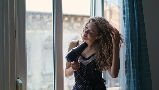 Woman using a hair dryer (heated styling) on curly hair—contrasting with heatless hair curler methods.