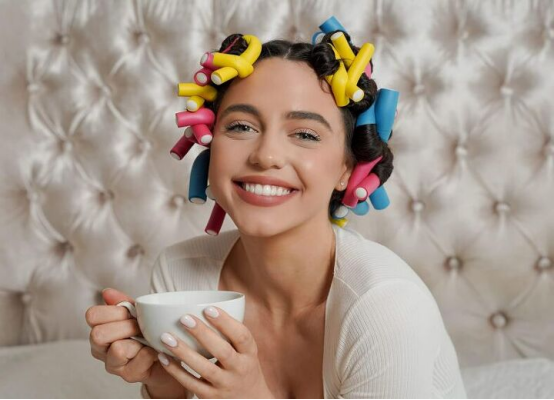 A woman with colorful foam heatless hair curlers in her hair, smiling with a cup, demonstrating the comfortable, at-home curling process.