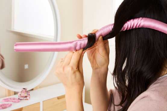 A woman wrapping hair around a pink satin heatless hair curler, demonstrating a simple, no-fuss curling step.